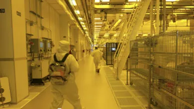 Workers in white hazmat suits walk down a sterile industrial corridor bathed in yellow light