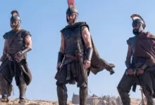 Three warriors in ancient Greek armor and crested helmets stand outdoors against a clear blue sky.