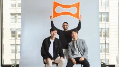 Three smiling men pose together in a photography studio, holding up a bright orange neon sign.