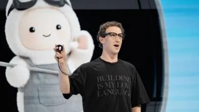 Mark Zuckerberg wearing a black t-shirt and glasses holding a small device on stage
