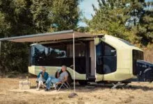 An older couple relaxing in camping chairs under the awning of a modern green and black RV.