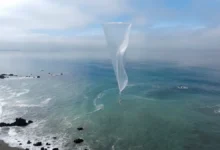 A white tapered balloon floating above a rocky coastline and blue ocean under a cloudy sky.