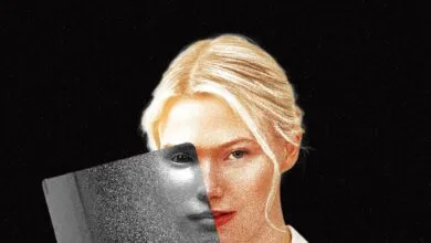 A blonde woman holding a glossy magazine partially over her face against a black textured background.