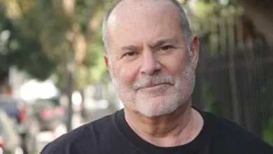 Portrait of an older Caucasian man with short grey hair and beard, wearing a black t-shirt outdoors.
