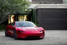 A sleek red Tesla Roadster parked in a gravel driveway in front of a modern home with a dark garage door.