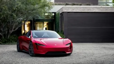 A sleek red Tesla Roadster parked in a gravel driveway in front of a modern home with a dark garage door.