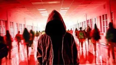 A hooded figure stands in the center of a crowded, red-tinted school hallway with blurred students.