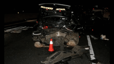 Severely damaged Highway Patrol vehicle with popped trunk and scattered debris on a dark highway at night.