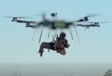 A person hanging suspended from a large multi-rotor drone flying against a pale sky.