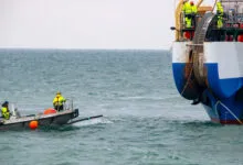 Workers on a small boat guide a thick undersea fiber optic cable from a large cable-laying ship.
