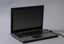 A dark gray Chromebook laptop sits open on a neutral gray surface with a charging cable plugged in.