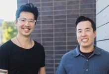 Two smiling men standing outdoors against a brick wall, wearing casual shirts.
