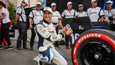 IndyCar driver Alex Palou smiling while kneeling beside his racing car with team members.