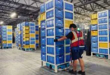 An Amazon worker guides a mobile robot carrying a tall stack of blue and yellow storage bins.