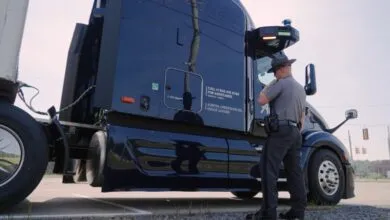 State trooper inspecting a dark blue Aurora self-driving semi-truck outdoors.