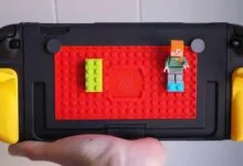 Hand holds a black handheld gaming console with a red LEGO baseplate on the back, featuring a green brick and a Minecraft Alex minifigure.