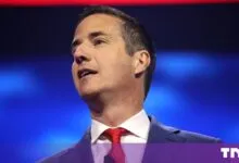 Bernie Moreno speaking on stage wearing a blue suit and red tie against a blue and red backdrop