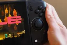 Close-up of a hand holding a black gaming handheld console with vibrant gameplay on screen.