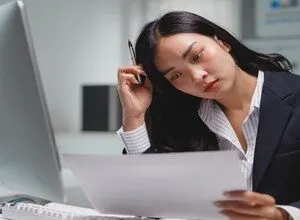 An Asian businesswoman looking stressed while reviewing documents at her office desk.