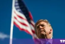 Donald Trump looking upward with an American flag waving in the background against a blue sky.