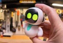 A hand holds a small white and blue desktop robot toy with glowing green digital eyes.