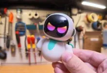 A person holds a small, cute white and teal robot toy with glowing pink eyes against a workshop background.