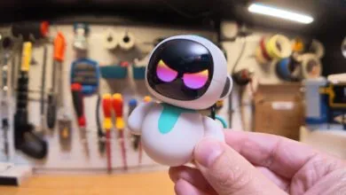 A person holds a small, cute white and teal robot toy with glowing pink eyes against a workshop background.