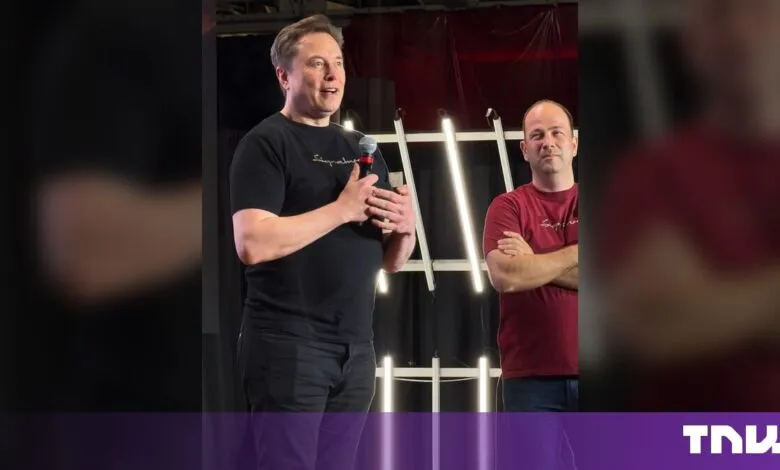 Elon Musk speaks into a microphone on stage while wearing a black t-shirt next to another man.