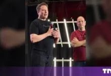 Elon Musk speaks into a microphone on stage wearing a black t-shirt next to another man.