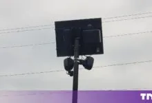 Flock dual camera unit mounted on a pole with a solar panel against a cloudy sky.