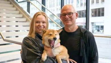 Sara and Tomas smiling alongside a yawning Shiba Inu dog indoors near a staircase.