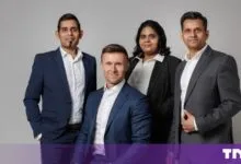 Four diverse business professionals posing together in dark suits against a plain grey backdrop.