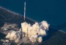 A rocket launches from a coastal pad, leaving a massive plume of white smoke and flying over a beach and ocean.