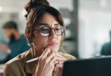 A businesswoman wearing round glasses thoughtfully focuses on her laptop screen in an office.