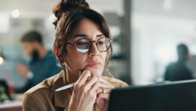 A businesswoman wearing round glasses thoughtfully focuses on her laptop screen in an office.