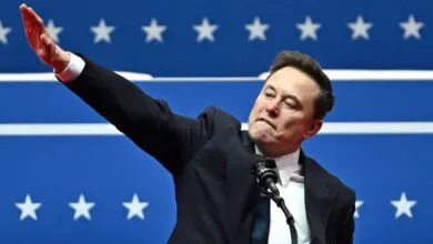 Elon Musk raises his arm while speaking at a political rally with a blue background featuring white stars.