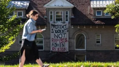 A student walks past a university building displaying a protest banner reading Protect Students Not Rapists