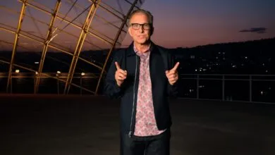 Greg Joswiak smiling and pointing fingers outward on an outdoor rooftop terrace at dusk.