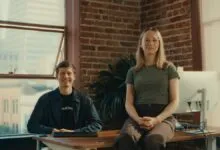 Two young professionals smiling in a modern brick-walled office with a city view.
