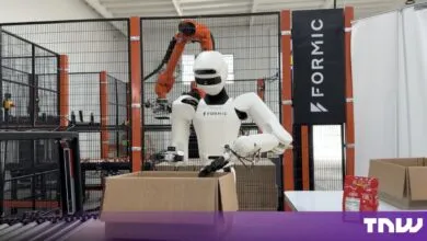A white humanoid robot packs a cardboard box inside a Formic factory cell with orange machinery.