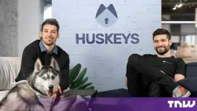 Two men posing in an office with a Husky dog in front of a painted brick wall featuring the Huskeys logo