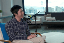 A man in a plaid shirt sits in a studio chair speaking into a podcast microphone.