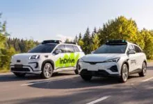 Two white autonomous vehicles from inDrive and Pony.ai driving on a sunny paved highway with trees.