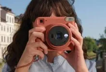 Young woman holding a terracotta-colored Instax Square camera to her face outdoors.