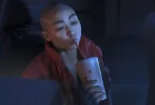 A bald character in a red jacket drinks from a cup with a red straw in a dimly lit sci-fi setting.