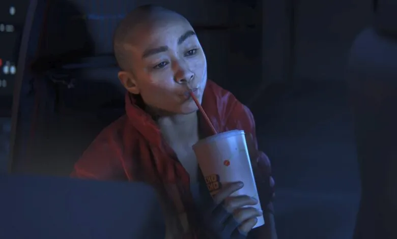 A bald character in a red jacket drinks from a cup with a red straw in a dimly lit sci-fi setting.