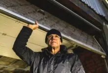 An older man in a winter jacket and beanie raises a metal garage door.
