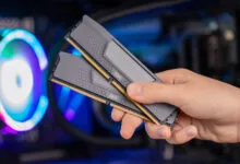 A person holds two black DDR5 RAM sticks in front of a glowing PC case.