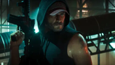 A bearded man in a hoodie and baseball cap holds a rifle in a dark, atmospheric setting.