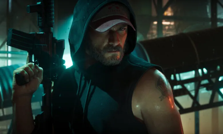 A bearded man in a hoodie and baseball cap holds a rifle in a dark, atmospheric setting.